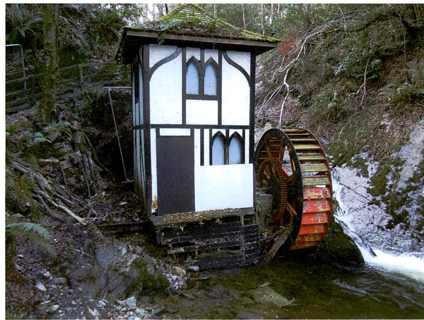 A photograph showing a white and black timber-framed wheelhouse with a large wooden waterwheel situated beside a stream in a wooded area.