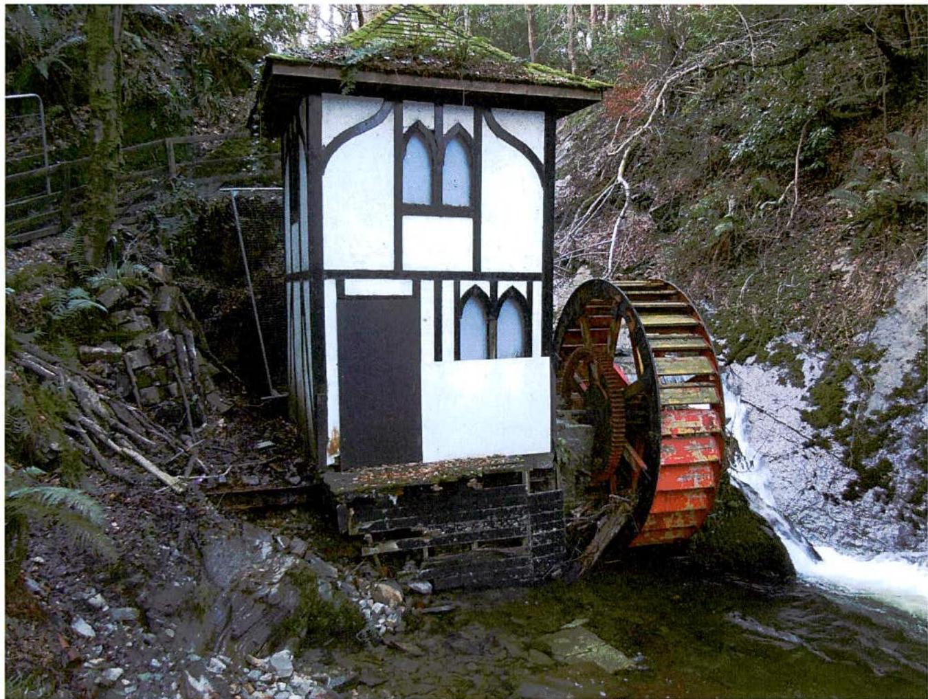 A photograph showing a small white and black timber-framed building with a large red water wheel situated beside a stream in a wooded area.