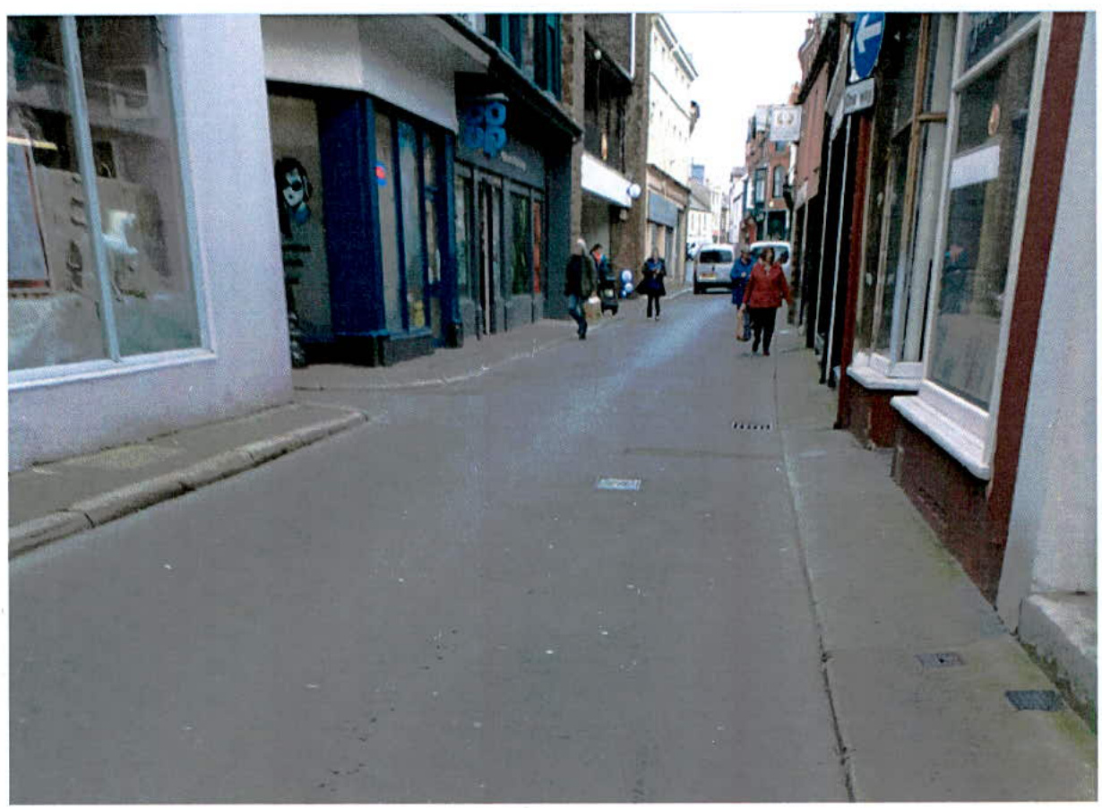 A street-level photograph showing a paved road in a town center with shop fronts on either side and pedestrians walking on the pavement.