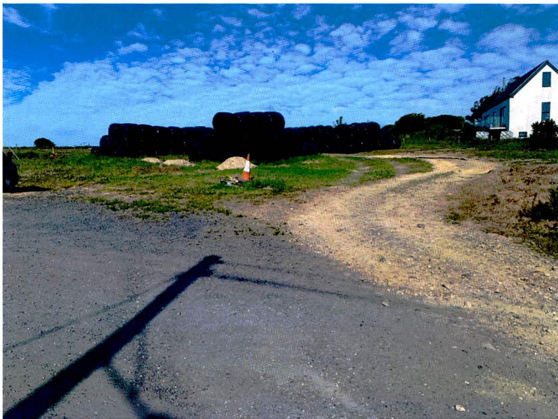 A photograph showing a rural site with a dirt track, stacked hay bales, and a white house in the background.