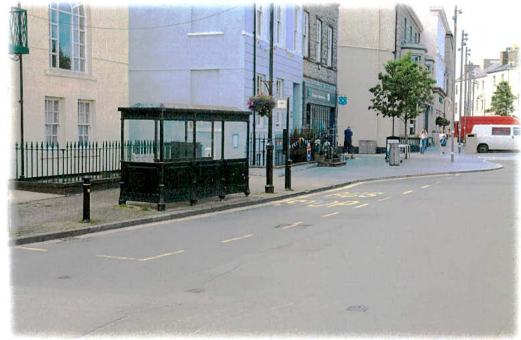 A photograph of a street scene featuring a black glass bus shelter on the pavement, with buildings and a road visible in the background.