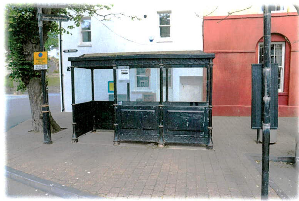 A photograph showing a black, ornate bus shelter on a paved sidewalk next to a tree and buildings.