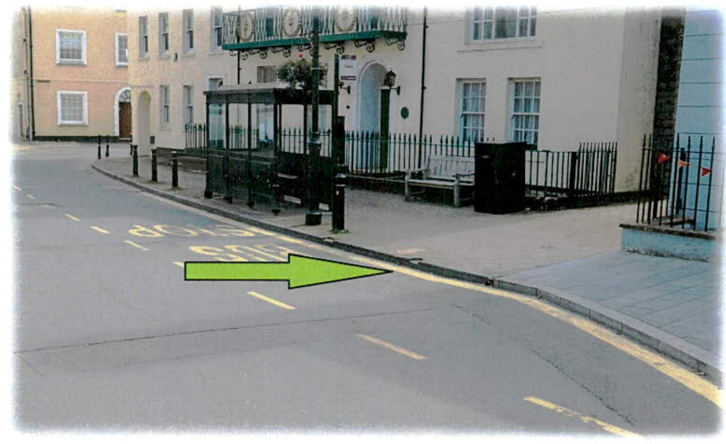 A street-level photograph showing an existing bus shelter on a sidewalk with a large green arrow superimposed pointing towards the curb area.