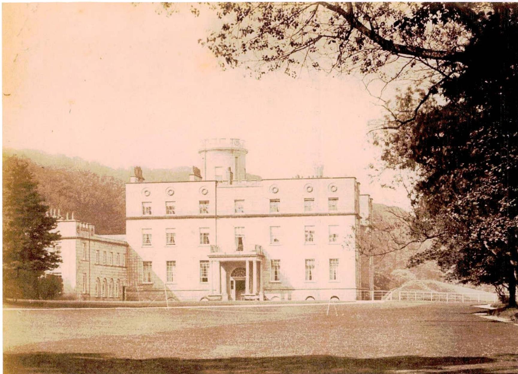 A sepia-toned historical photograph depicting a large, multi-story stately home or mansion with a central tower, set against a hilly backdrop.