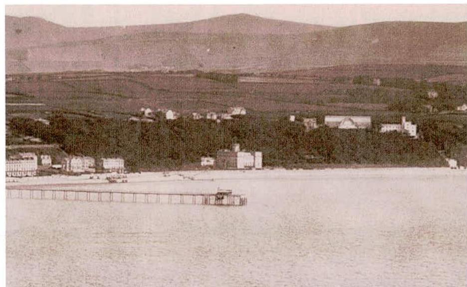 A vintage sepia-toned photograph depicting a coastal scene with a long pier extending into the water, a row of buildings along the shore, and hills in the background.