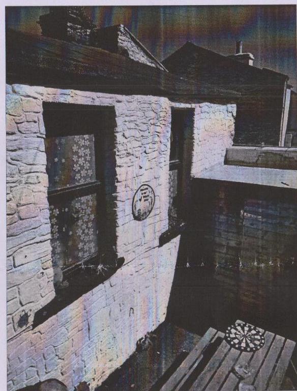 A grainy photograph showing the stone exterior wall of a building with a window and a circular plaque, next to a wooden deck with a dartboard.