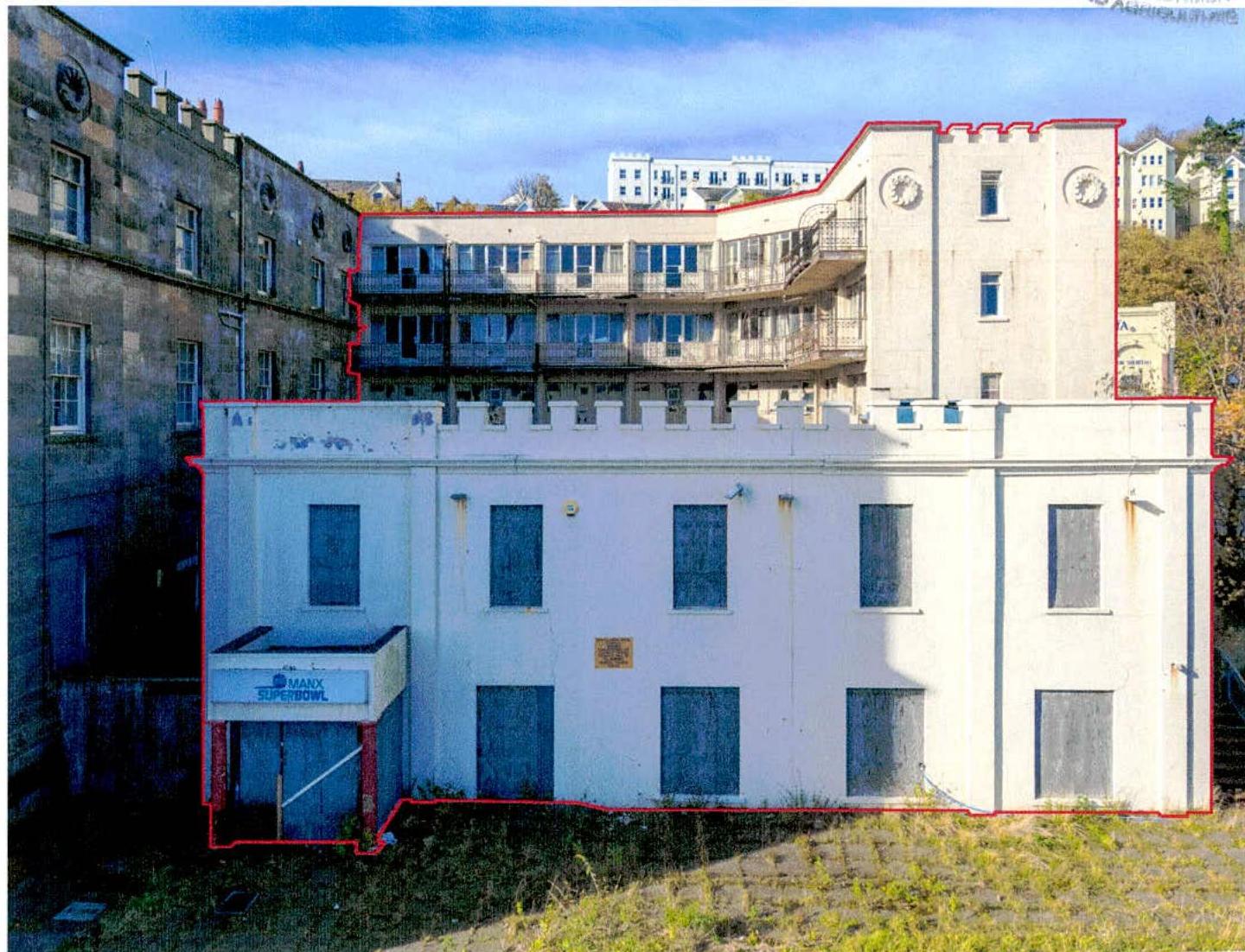 A photograph showing a large white commercial building with boarded windows and a 'Manx Superbowl' sign, outlined in red. Behind it is a curved, multi-story building with balconies, also appearing derelict.
