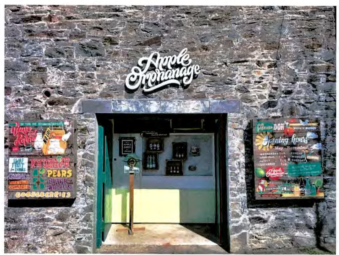 A photograph showing the exterior of a stone building with a sign reading 'Apple Orchannage' above a doorway, flanked by posters advertising produce and opening hours.