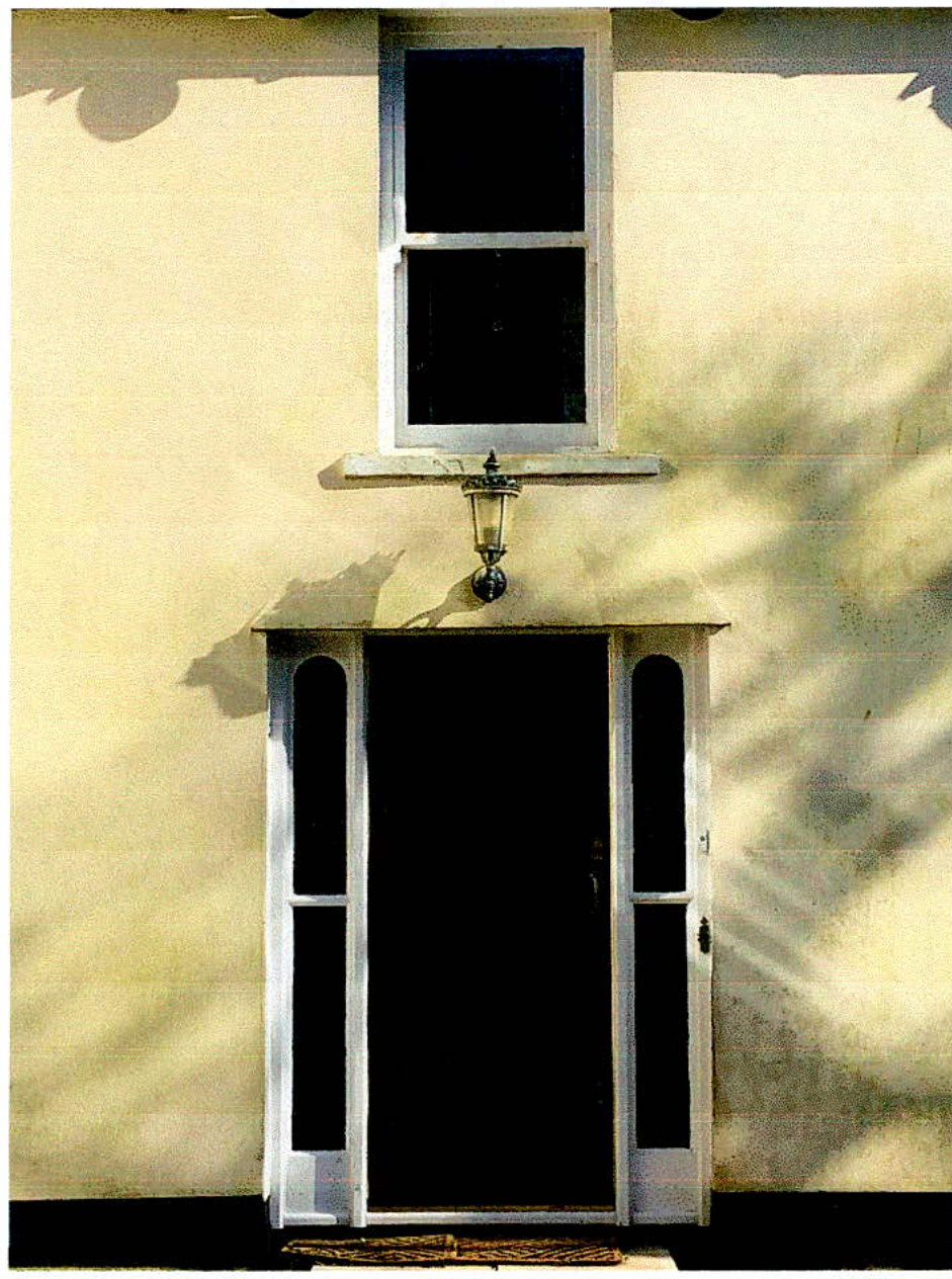 A photograph showing the front entrance of a building with a dark door, white sidelights, and a window above on a cream-colored wall.