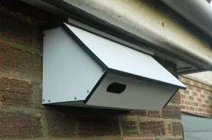A close-up photograph showing a white dormer window structure attached to a brick wall, featuring a sloped roof and a small opening.