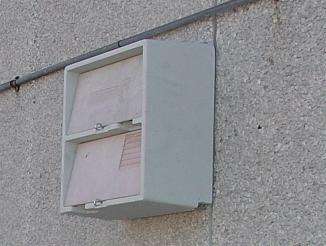 A close-up photograph of a grey utility box mounted on a roughcast exterior wall with its door open, revealing documents inside.