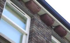 A close-up photograph of a building exterior showing a white window frame, brickwork, and terracotta guttering along the roofline.