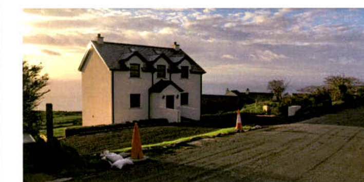 A photograph showing a white, two-story detached house with a paved driveway and construction cones in the foreground, set in a rural landscape.