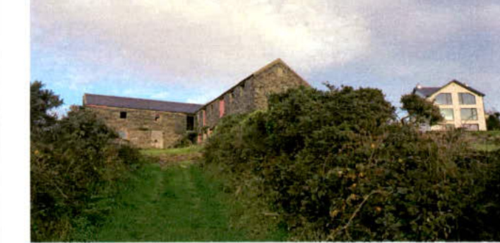 A photograph showing a large stone agricultural building and a modern white house situated in a rural, overgrown landscape.