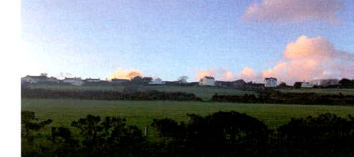 A landscape photograph showing a green field in the foreground with a row of houses visible on a hill in the distance under a cloudy sky.