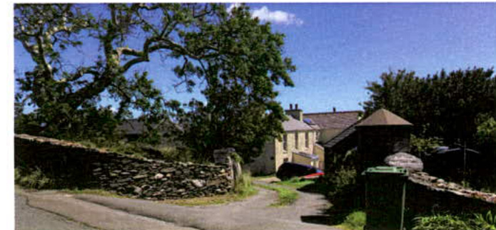A photograph showing a rural street scene with a stone boundary wall, driveway access, and a house partially obscured by trees.
