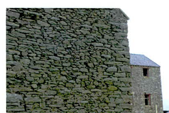 A close-up photograph of a rough stone wall and the corner of a stone building with a slate roof and window openings.