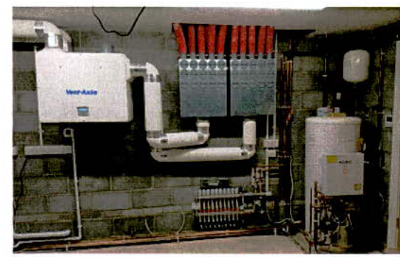 A photograph of a utility room interior featuring a ventilation unit, electrical distribution board, and a hot water cylinder.