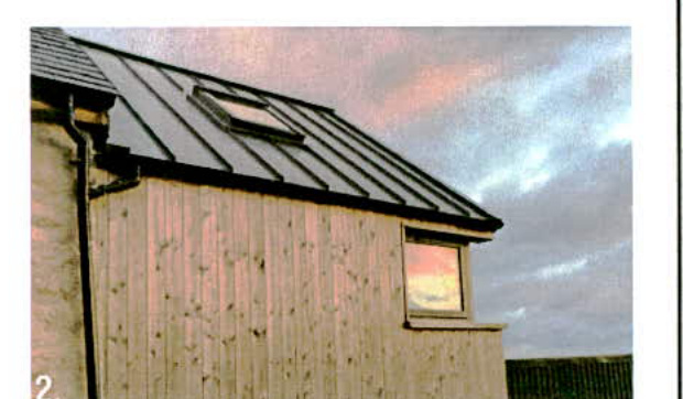 A close-up photograph of a building exterior featuring vertical timber cladding, a dark pitched roof with a skylight, and a window against a cloudy sky.