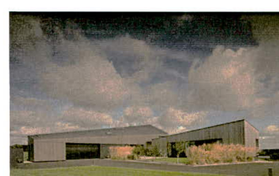 A grainy exterior view of a modern, single-story detached building with dark cladding and large windows, set against a cloudy sky with landscaping in the foreground.
