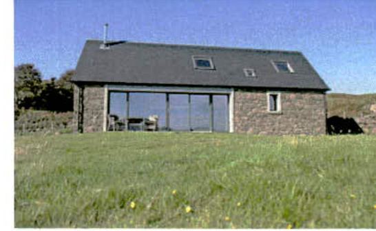 A photograph of a detached stone bungalow with a pitched roof and large glass doors, situated in a grassy field.