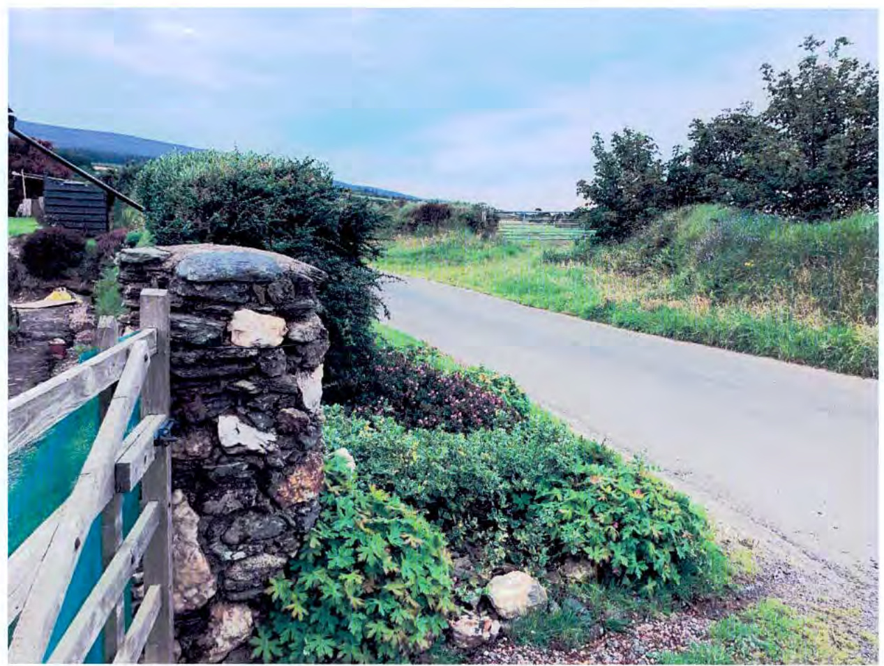A photograph showing a rural road scene with a stone wall and wooden gate in the foreground, looking towards a field and hills in the distance.