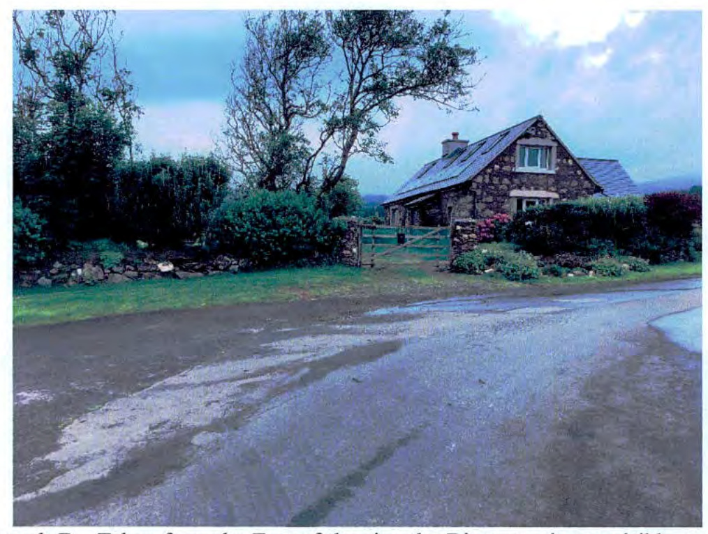A photograph of a stone detached house with solar panels on the roof, viewed from the road with a driveway and gate.