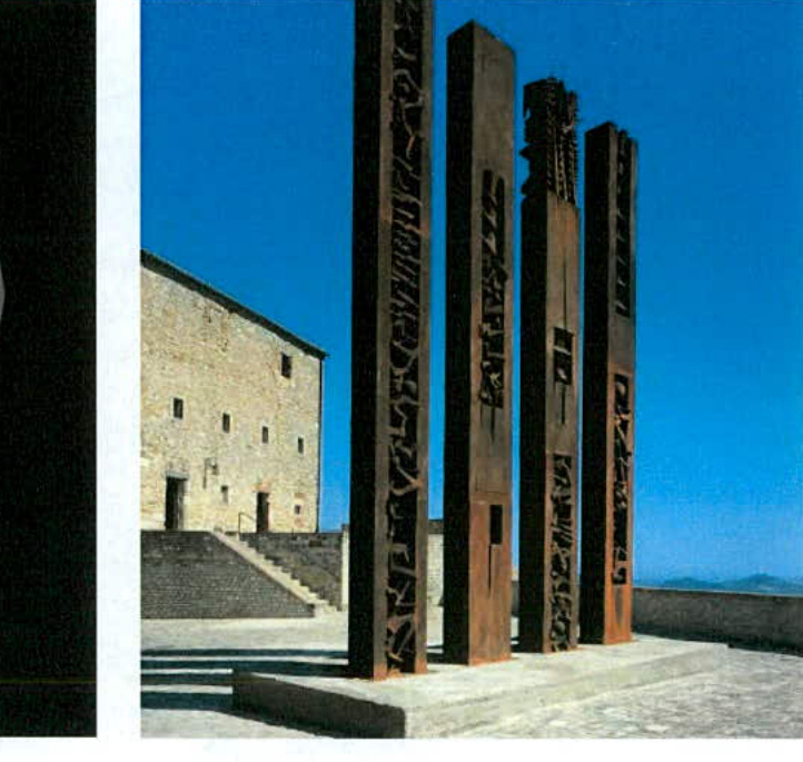 A photograph showing four tall, rusted metal feature totems standing on a concrete plinth with a stone building in the background.