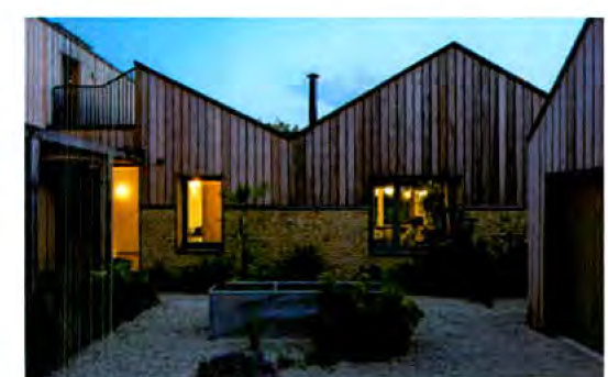 A photograph showing the exterior of a modern residential property with vertical timber cladding and stone walls, featuring a gravel courtyard and illuminated windows at dusk.