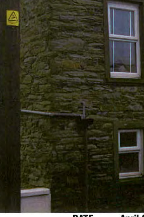 A close-up photograph of a rough stone wall featuring two white-framed windows and a yellow utility warning sign on a wooden post.