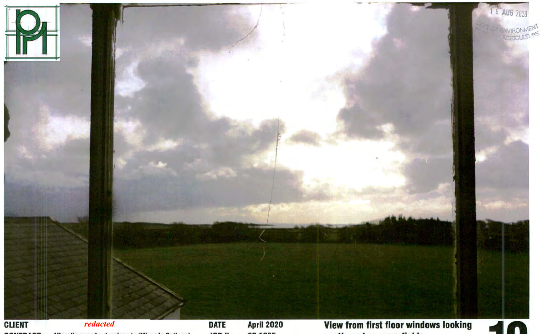 A photograph taken from inside a building looking out through a window frame at a green field and distant trees under a cloudy sky.