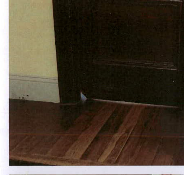 A close-up photograph showing the bottom of a dark wooden door meeting a wooden floor, documenting the existing interior condition.