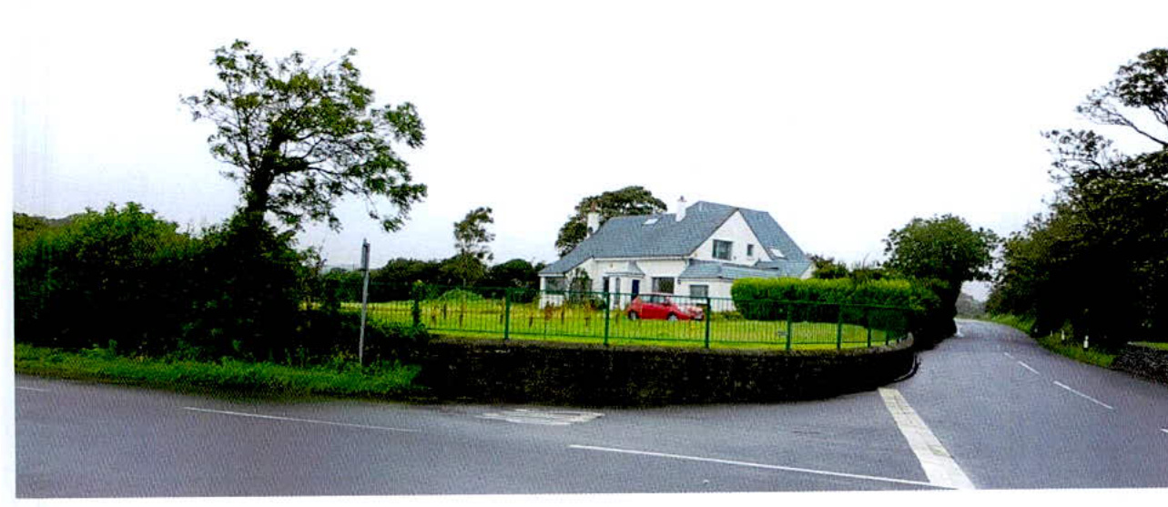 A street-level photograph showing a white detached house with a grey roof situated at a road junction, featuring a red car parked on the lawn and a green fence along the boundary.