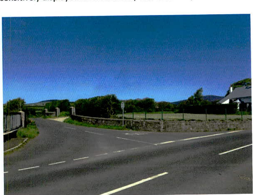 A photograph showing a rural road junction with a stone wall, green fencing, and a house visible in the background under a clear blue sky.