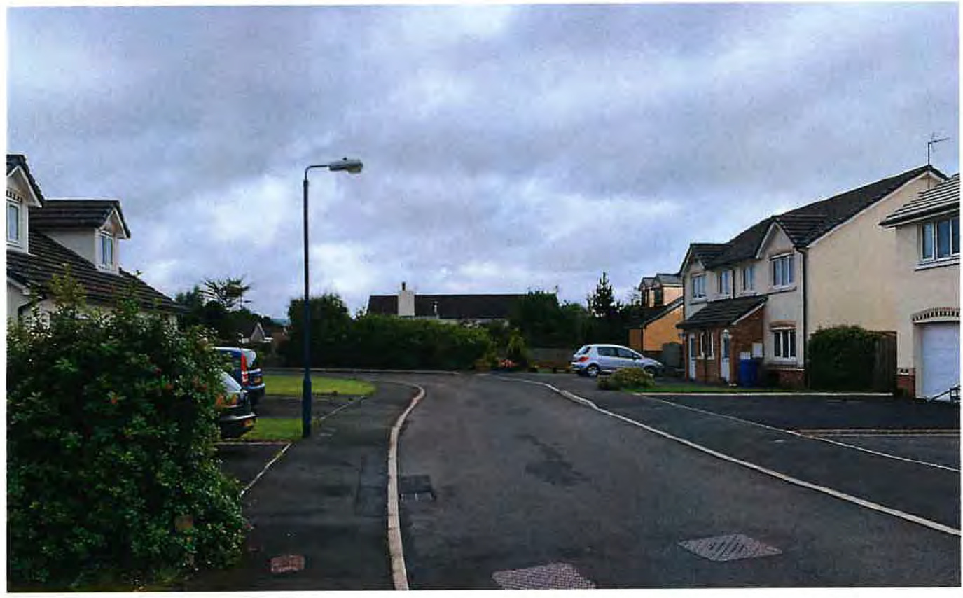 A street-level photograph showing a residential road with detached houses, parked cars, and a street lamp under a cloudy sky.