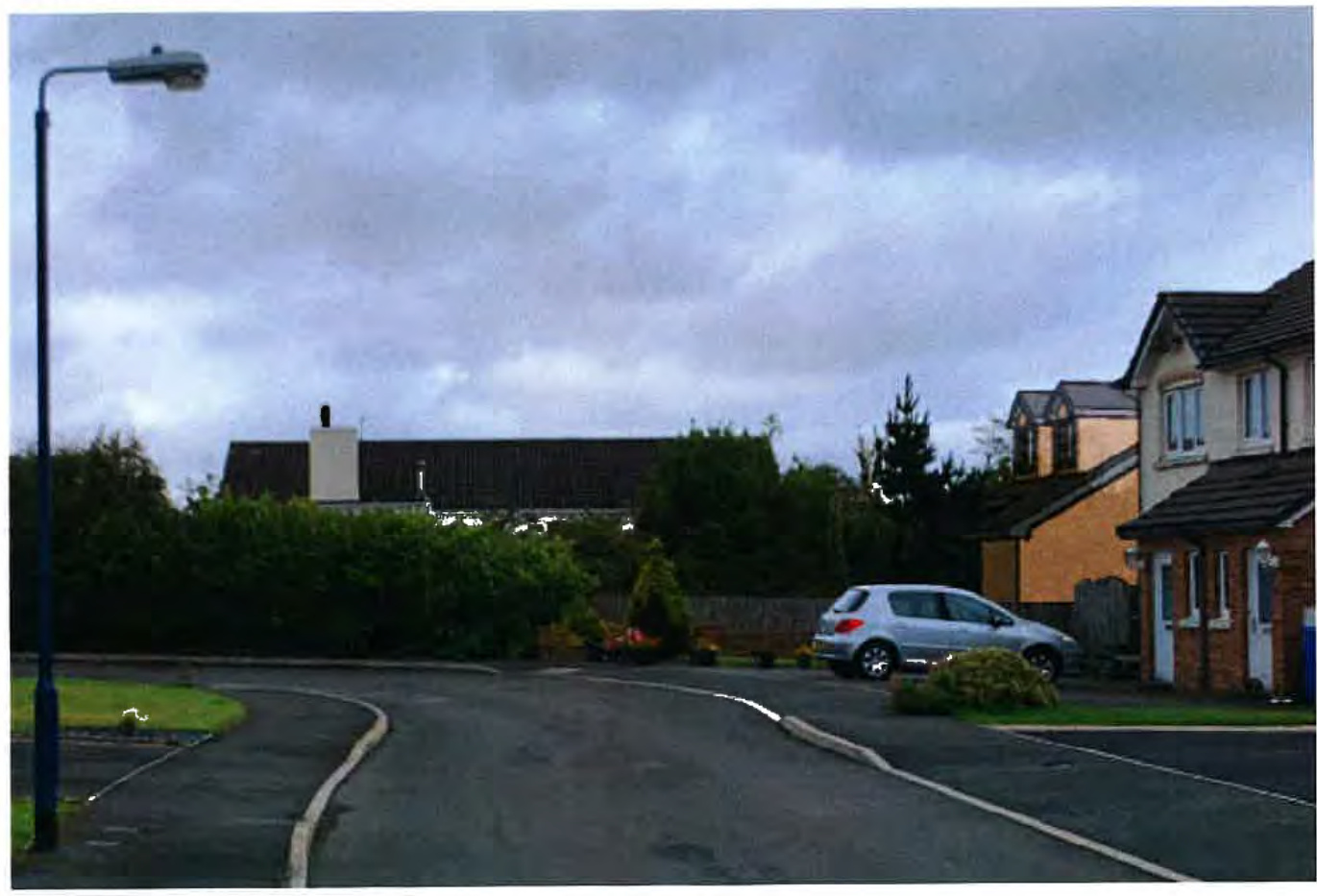 A street-level photograph showing a residential road curving to the left with a parked car and neighboring houses under a cloudy sky.