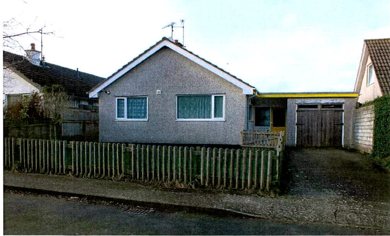 A photograph showing the exterior of a single-story detached bungalow with pebbledash rendering, a wooden front fence, and an attached garage with a driveway.