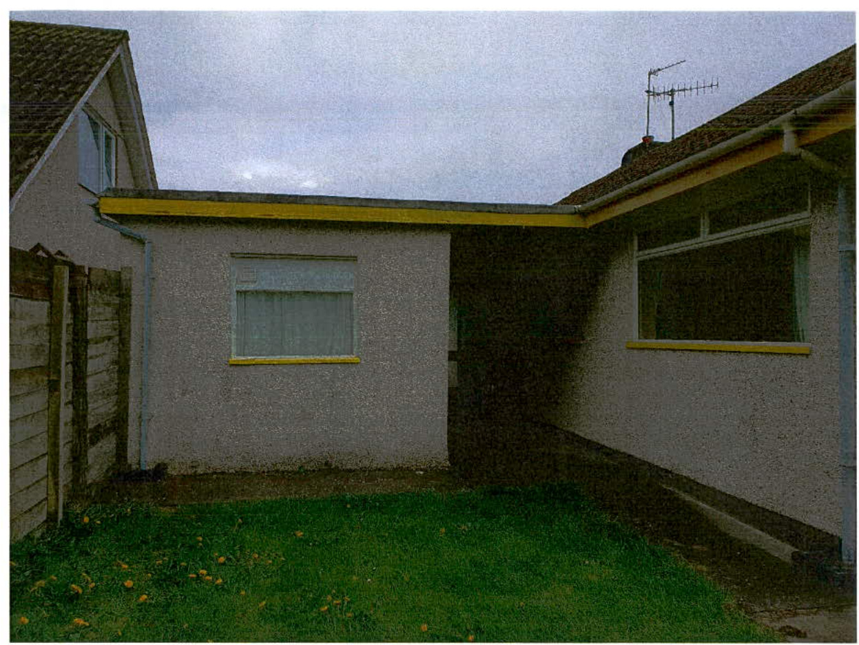 A photograph showing the side elevation of a single-story white rendered building with yellow window trim, situated next to a wooden fence and grassy area.