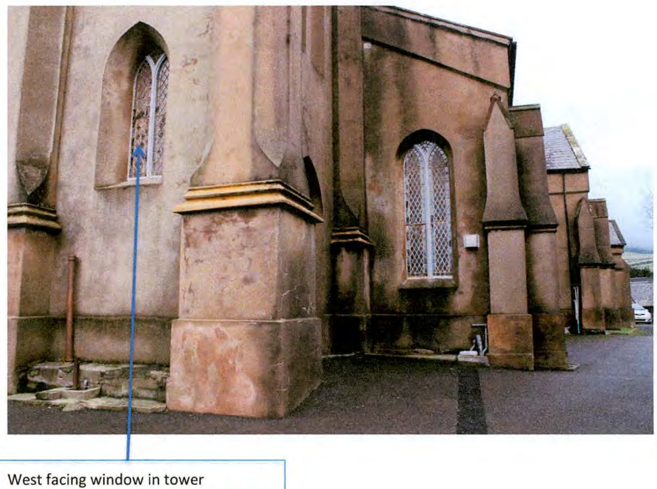 A photograph showing the exterior stone walls of a church, specifically highlighting a tall arched window in the tower with a blue arrow and caption.
