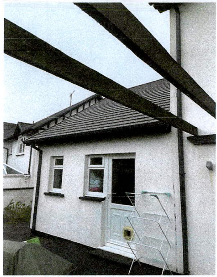 A grainy photograph showing the side or rear elevation of a white single-story bungalow with a tiled roof and large timber beams in the foreground.