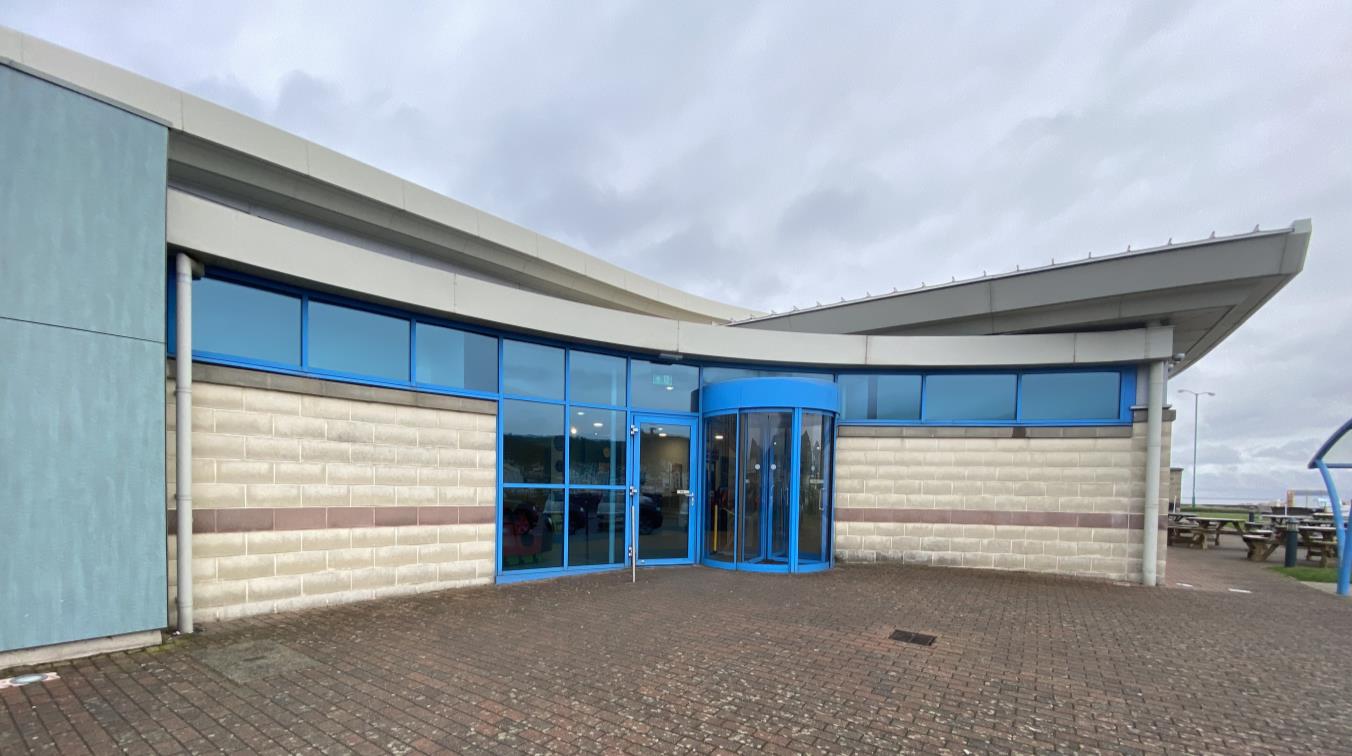 A photograph of a modern commercial building exterior featuring a blue-framed glass entrance with a revolving door. The building has light brick cladding and a paved forecourt area with picnic tables in the background.