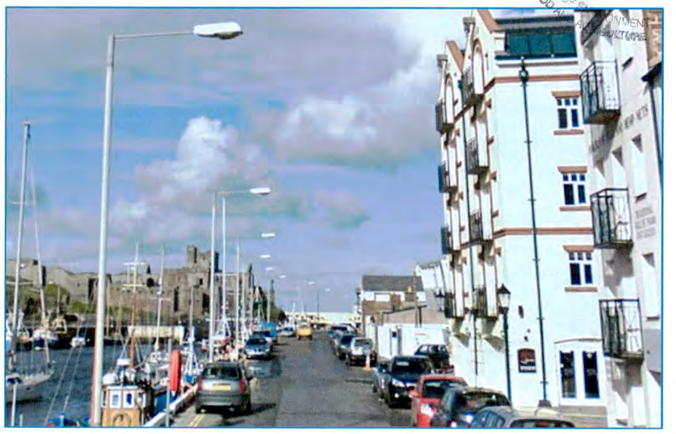A street scene in Peel showing a harbor with boats on the left and white multi-story buildings on the right, with Peel Castle visible in the background.