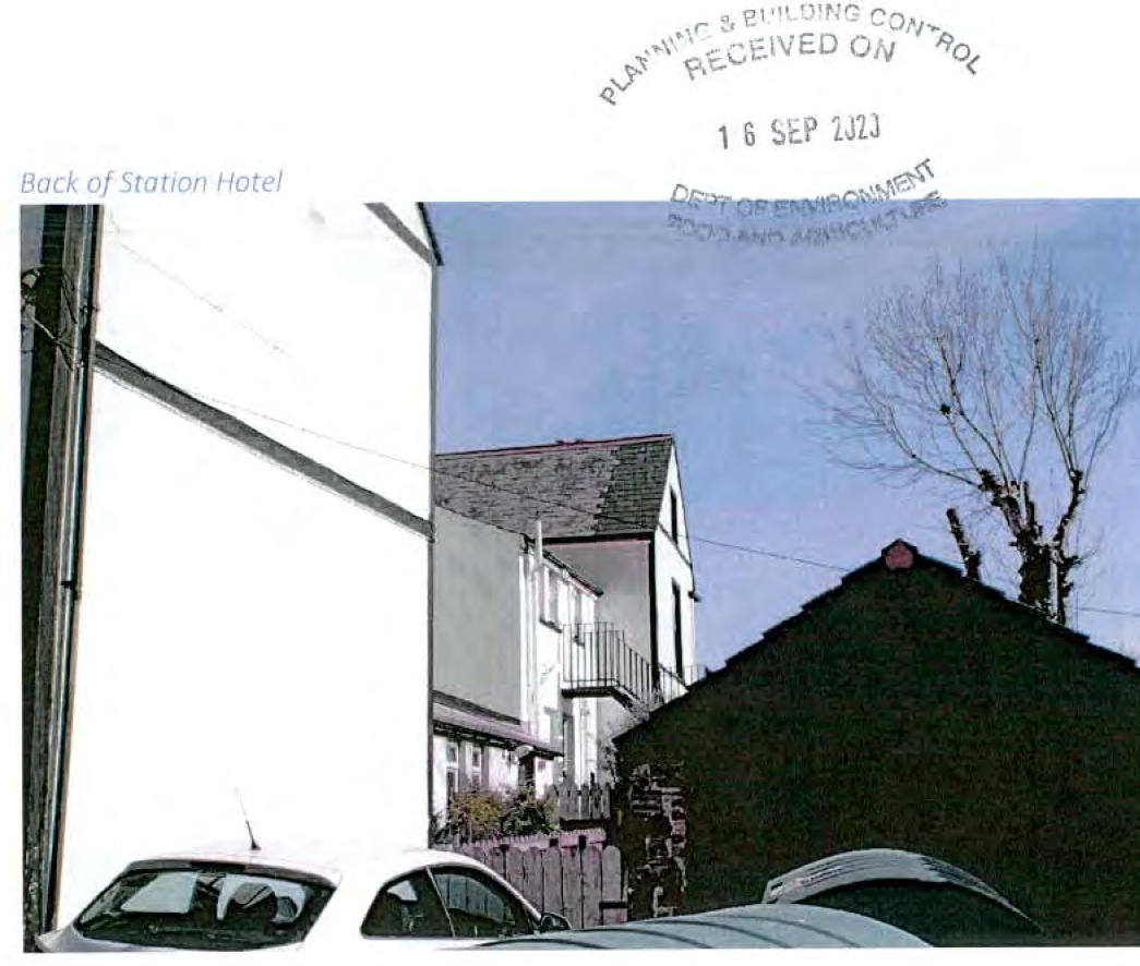 A photograph showing the rear of the Station Hotel and adjacent residential properties, including a white house and a dark outbuilding, with cars parked in the foreground.