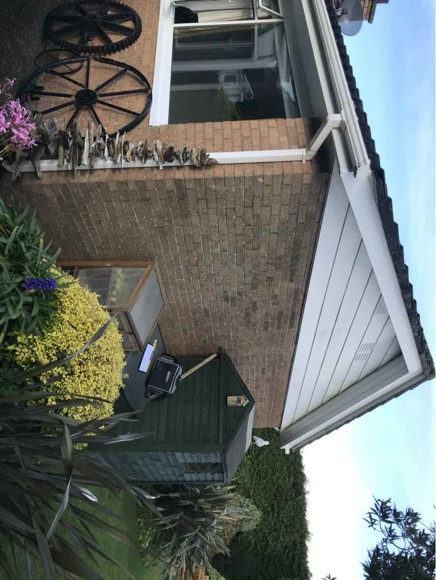 A rotated photograph showing the brick exterior of a house with a window, decorative wheels, and a garden area containing a shed and greenhouse.