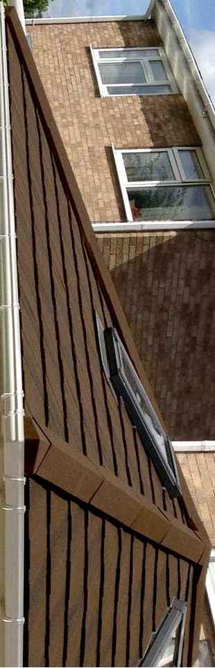 A low-angle photograph showing the junction of a brown tiled roof with skylights and an adjacent brick wall with white-framed windows.
