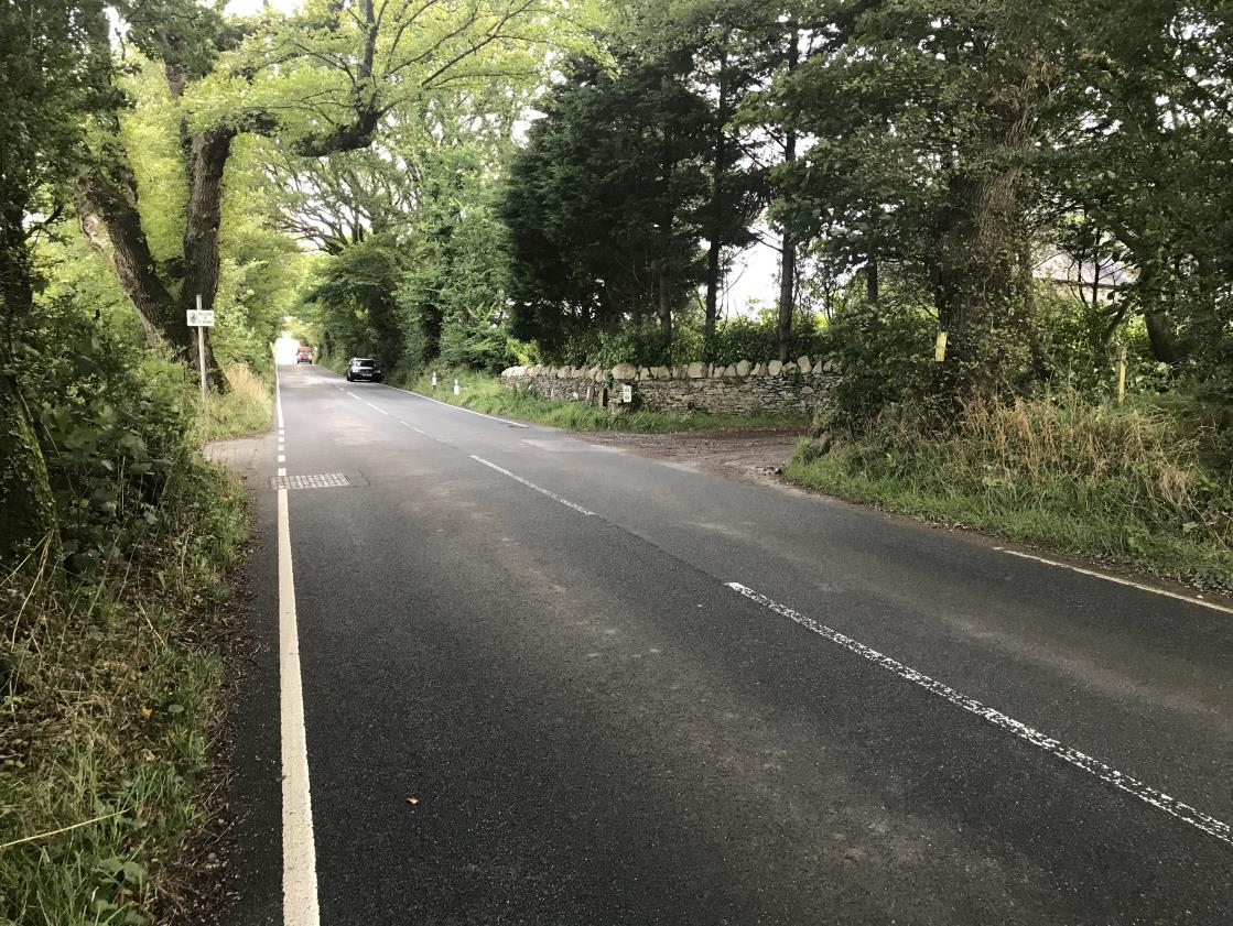 A photograph showing a paved rural road with trees and a stone wall on the right side, likely depicting the site of the proposed vehicular access.