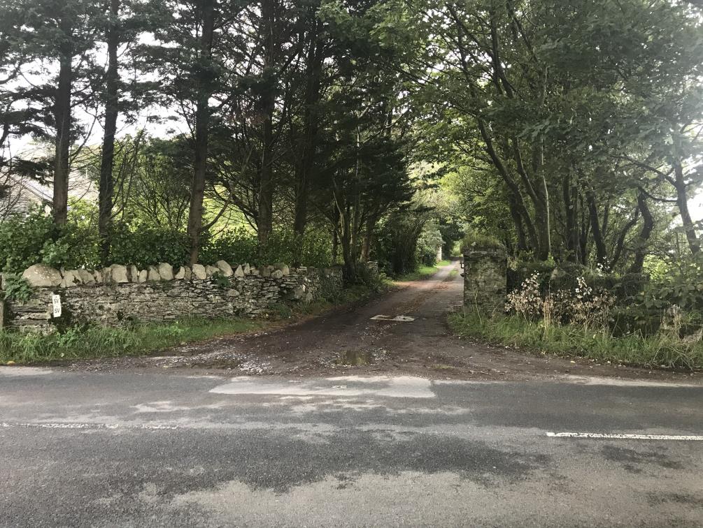 A photograph showing a rural road junction with a gravel driveway entrance flanked by stone walls and trees.
