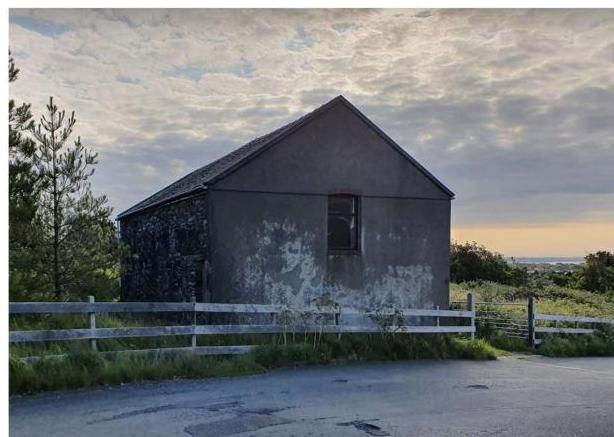 A photograph showing a weathered, single-story stone and rendered building with a pitched roof, situated behind a white fence in a rural coastal setting.