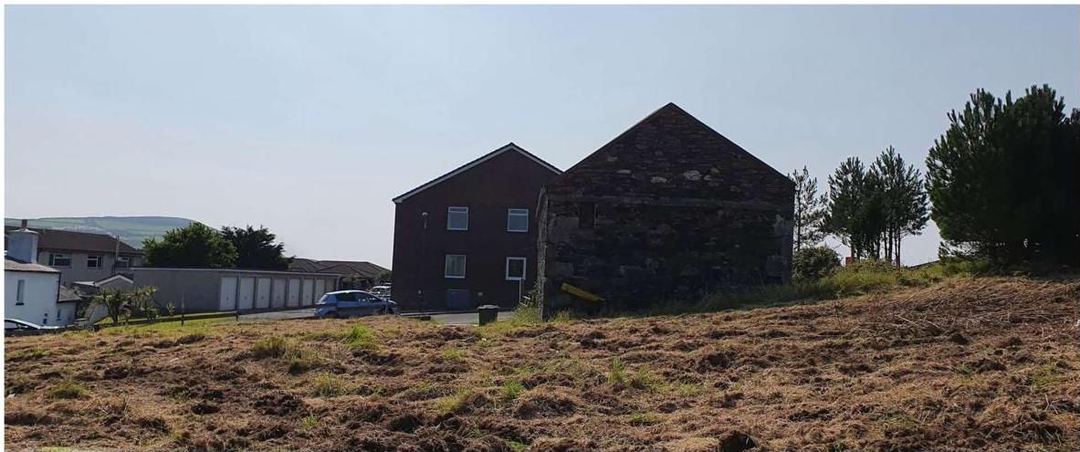 A photograph showing a grassy plot of land in the foreground with existing buildings, including a stone barn and a modern house, in the background.