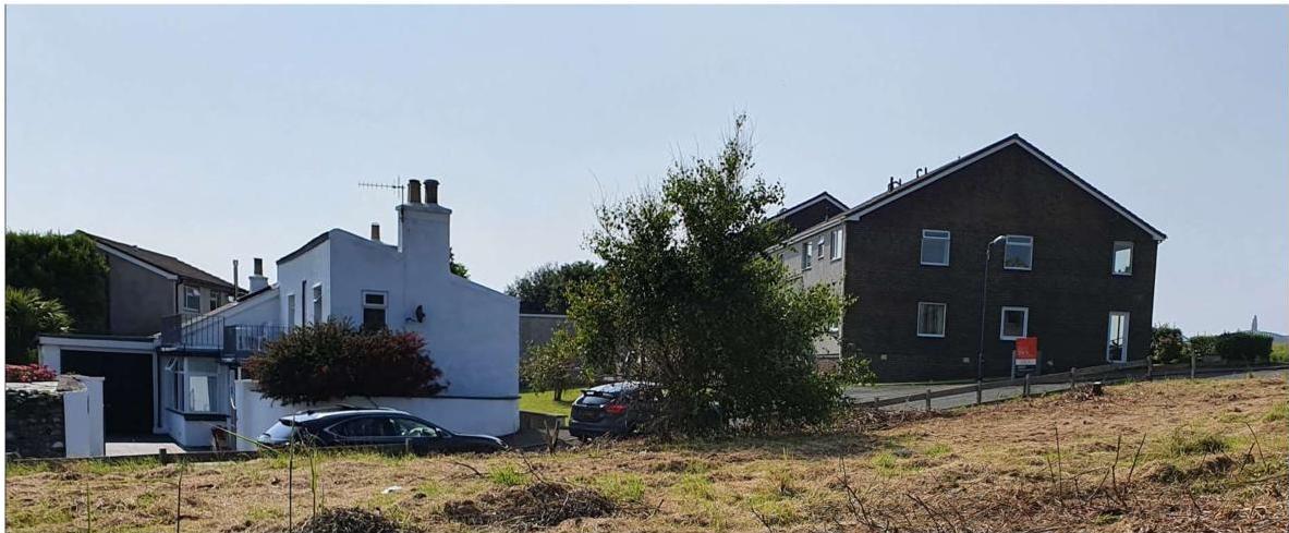 A photograph showing a street view of existing residential properties, including a white house and a dark brick house, with a grassy foreground area.